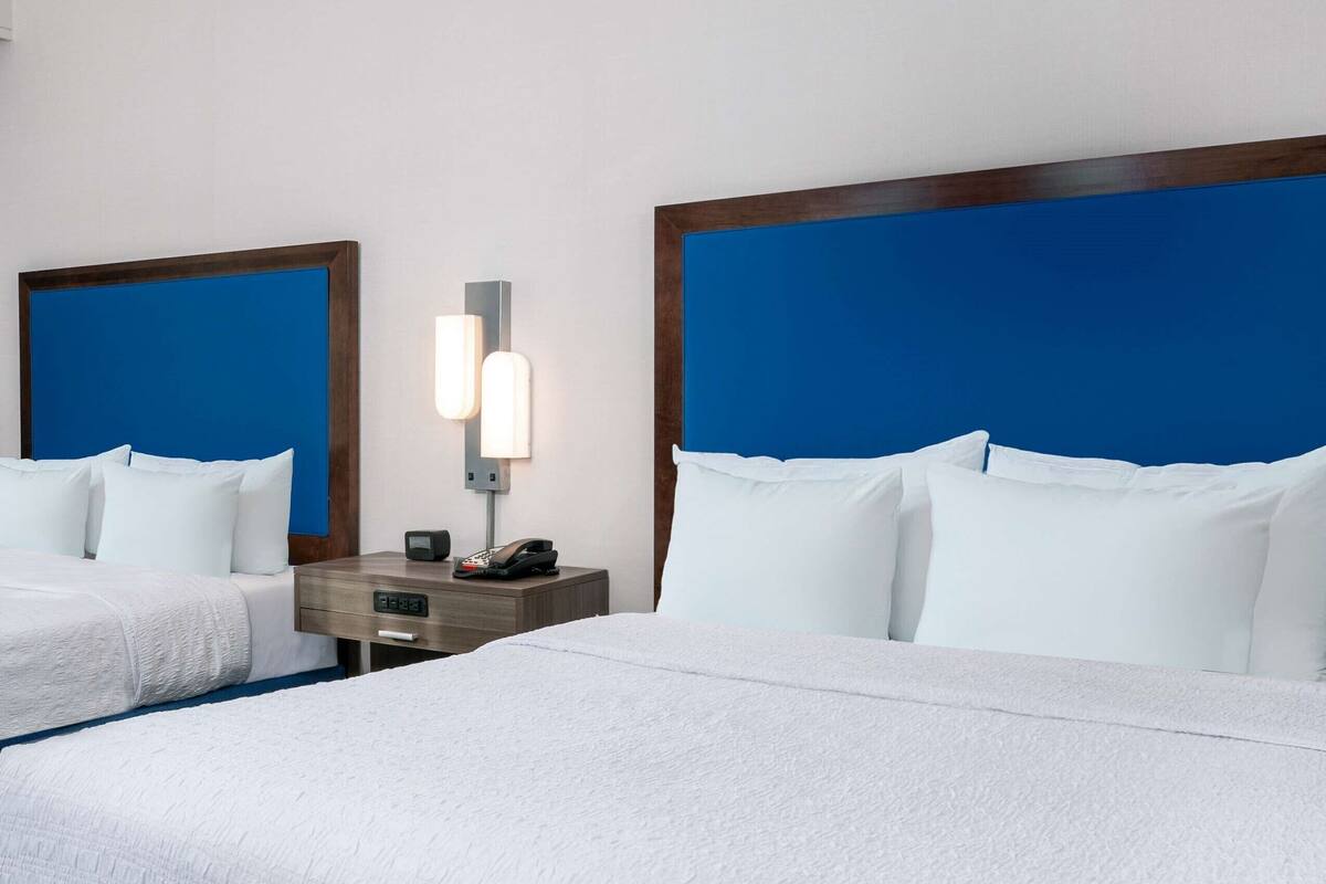 A hotel room with two beds, a nightstand with a phone, and a wall-mounted light.