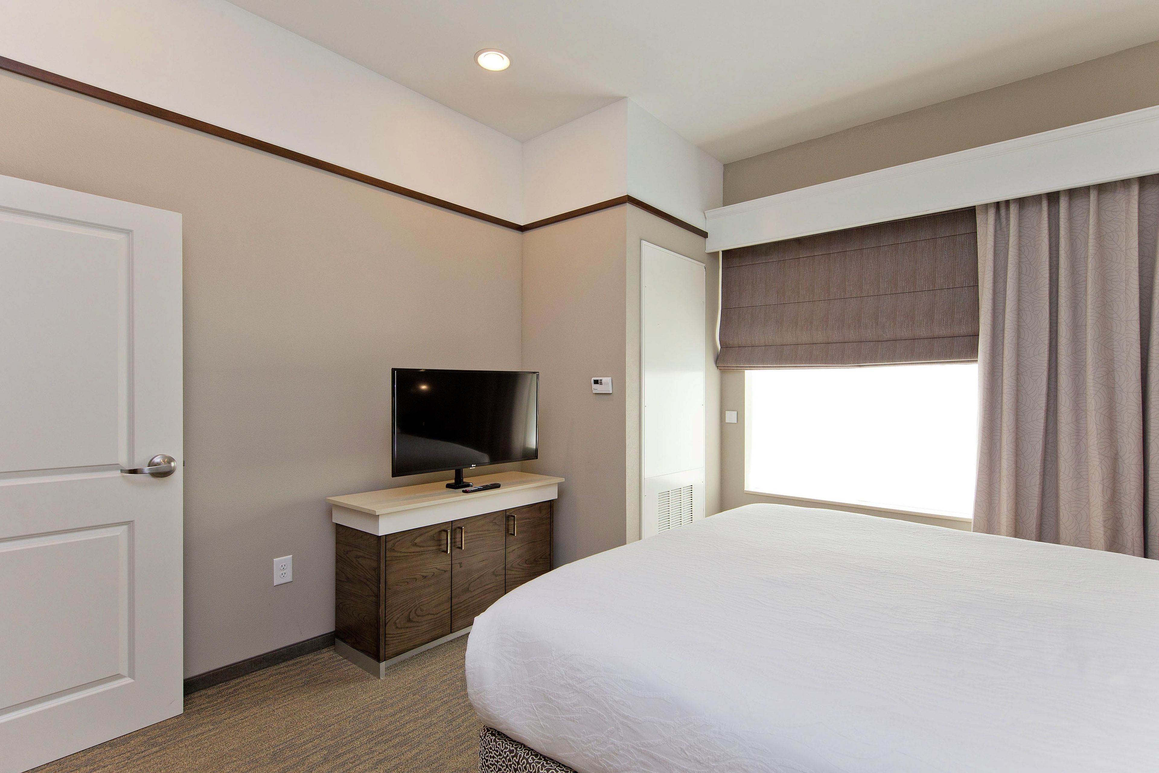 Quarto de hotel com cama, televisão em suporte, guarda-roupa e janela com cortinas.