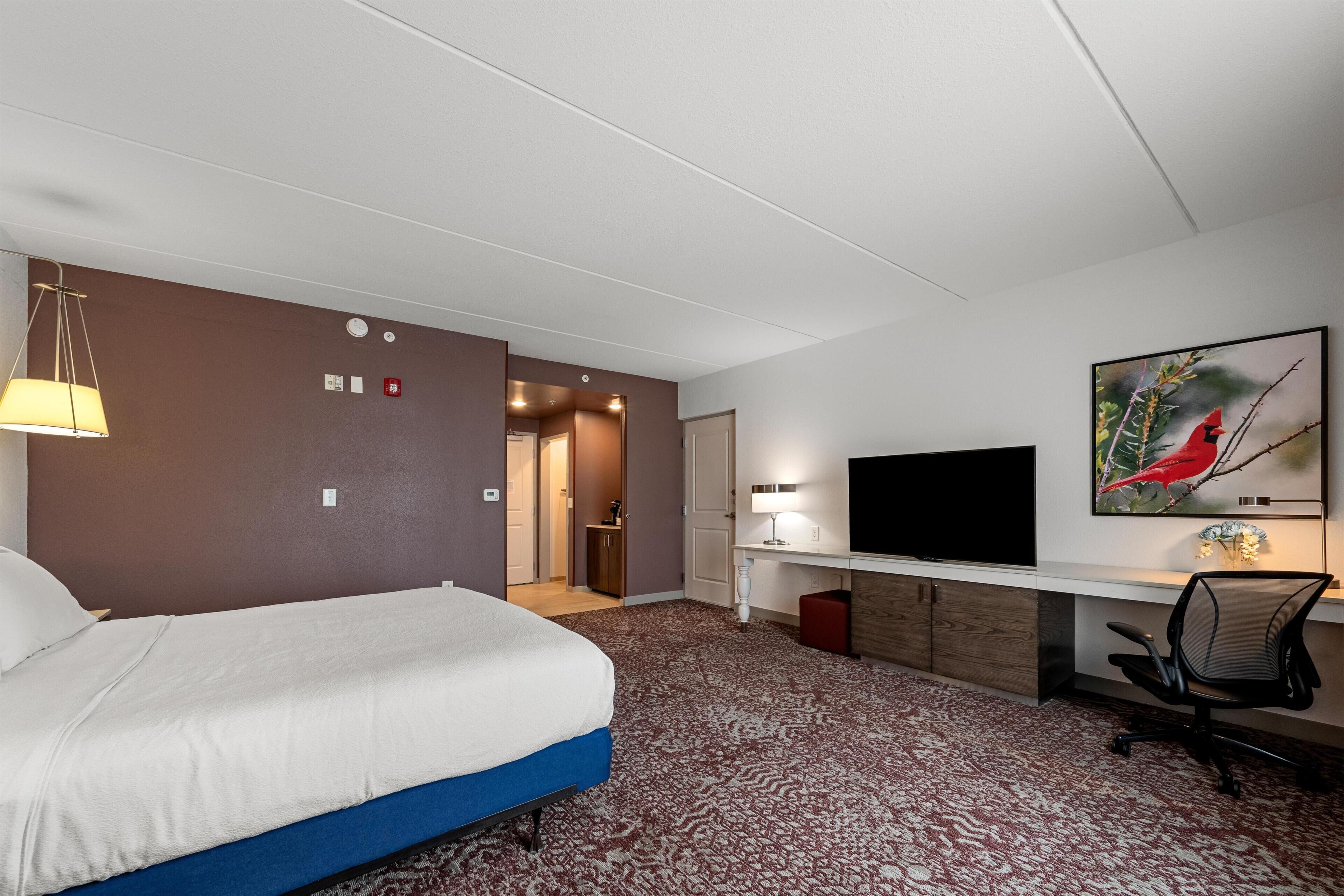 A hotel room with a bed, desk, chair, TV, and artwork.