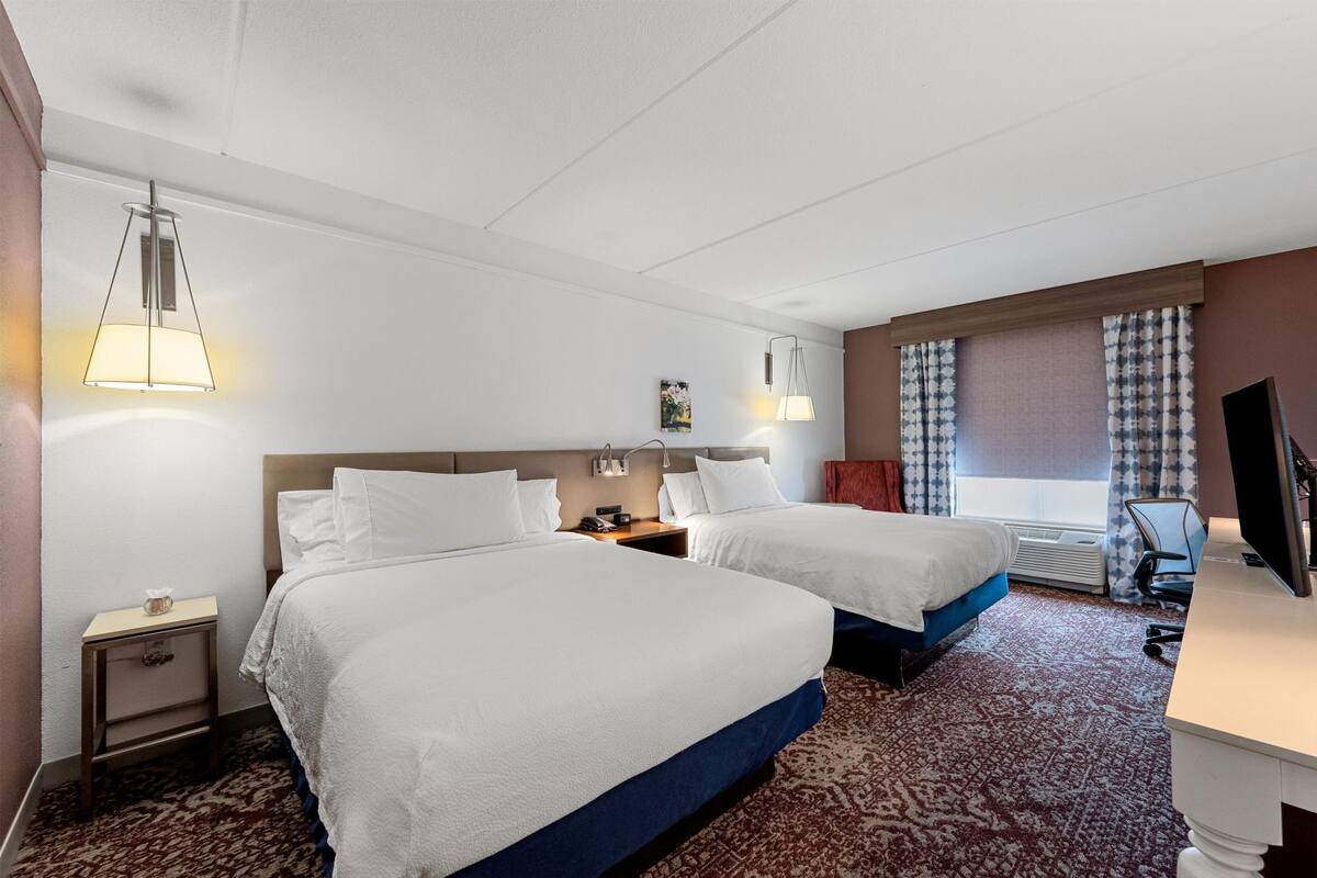 Ein Hotelzimmer mit zwei Betten, einem Schreibtisch, einem Stuhl, einem Fernseher und einem Fenster mit Vorhängen.