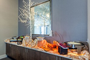 A buffet setup with various food items, a decorative tree, and a window with natural light. - Hilton Garden Inn Martinsburg (Martinsburg)