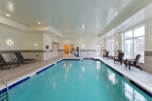 Una piscina interior con agua cristalina, rodeada de sillones de descanso y una tumbona, con vistas al exterior.