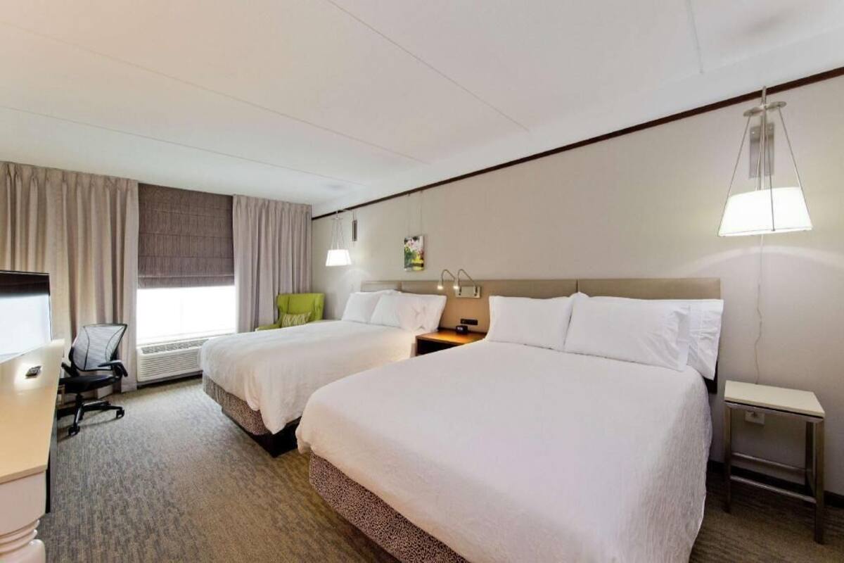 Ein Hotelzimmer mit zwei Betten, einem Schreibtisch, einem Stuhl, einer Lampe und einem fenster mit Vorhängen.