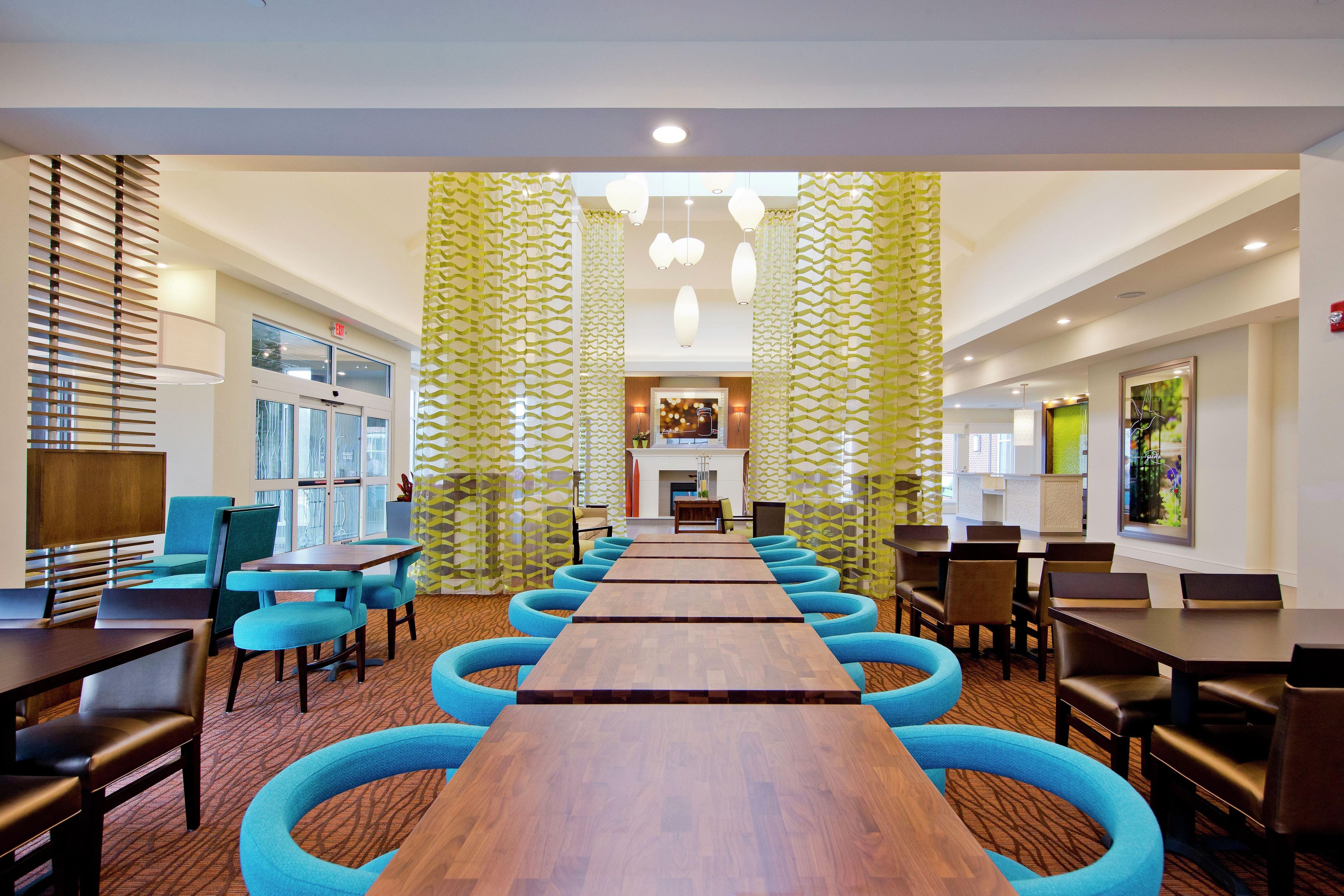 A hotel conference room with a long wooden table, blue cushioned chairs, and patterned curtains.