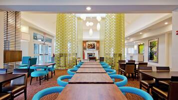 A hotel conference room with a long wooden table, blue cushioned chairs, and patterned curtains.