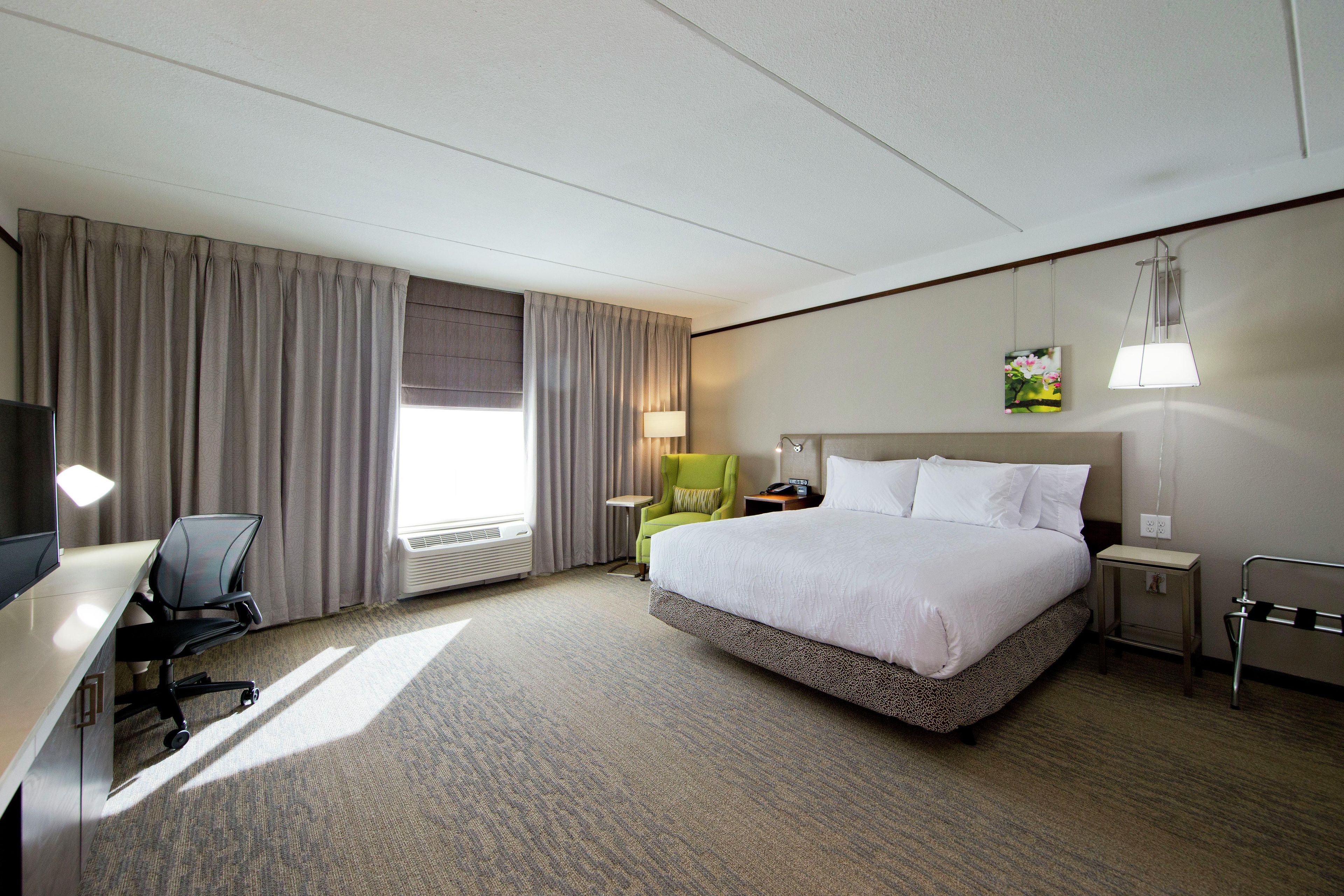 A hotel room with a bed, a desk, a chair, a lamp, and a painting on the wall.