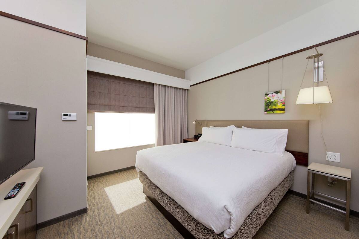 Ein Hotelzimmer mit einem großen Bett, einem Fernseher, einem Nachttisch, einer Lampe und einem Fenster mit Vorhängen.
