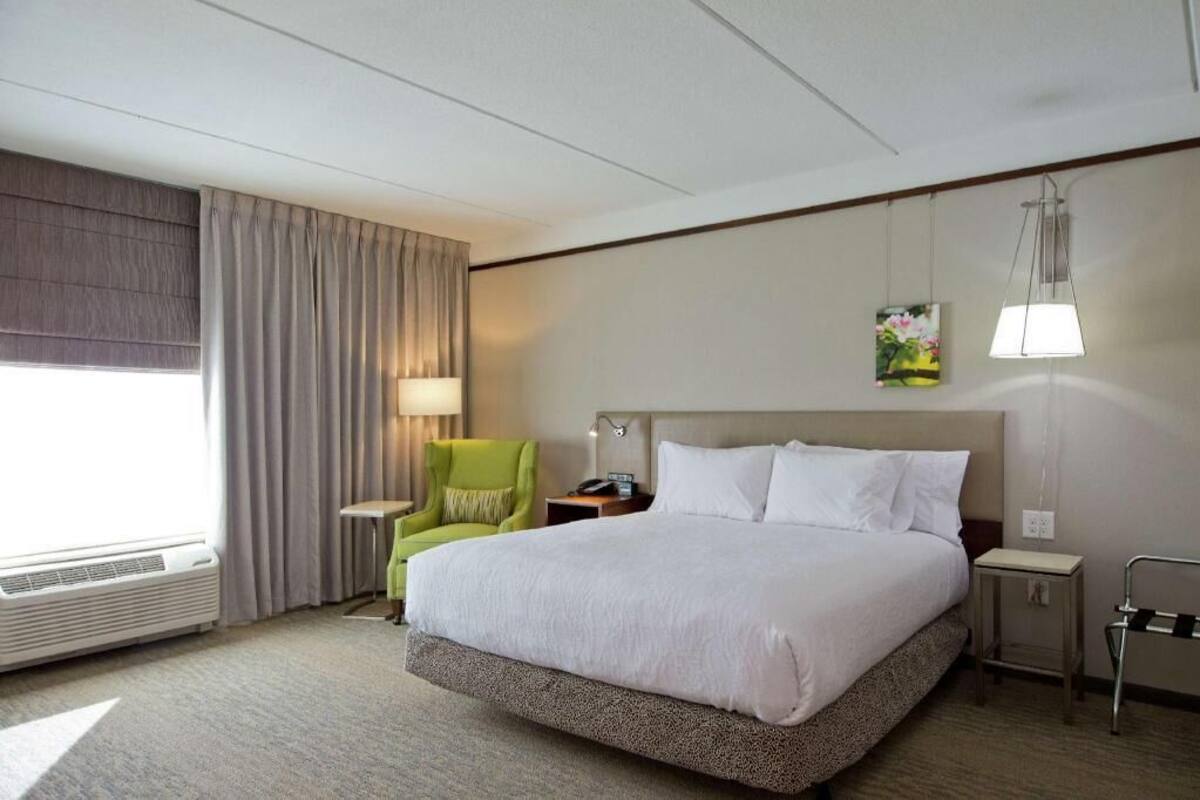 Ein Hotelzimmer mit einem großen Bett, einem Sessel, einem Nachttisch, einer Lampe und einem Bild an der Wand.