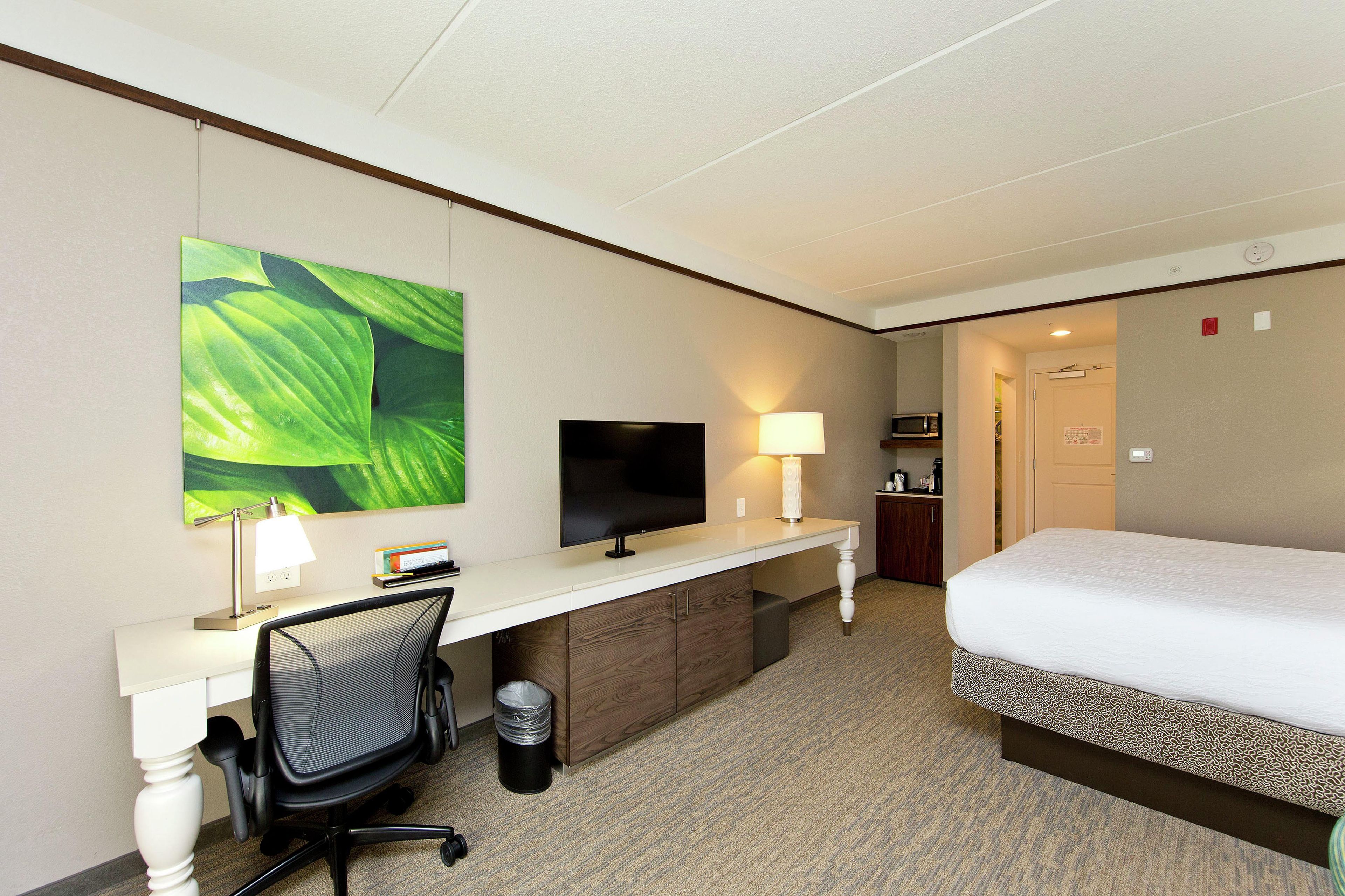 A hotel room with a bed, desk, chair, TV, lamp, trash can, and a microwave.