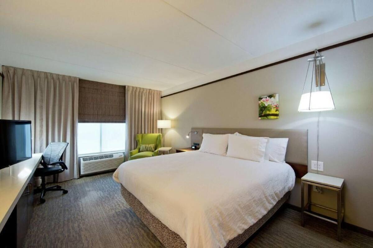 Ein Hotelzimmer mit einem großen Bett, einem Schreibtisch mit Stuhl, einem Fernseher, einer Lampe und einem Fenster mit Vorhängen.