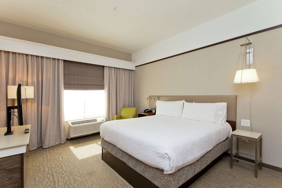 Ein Hotelzimmer mit einem großen Bett, einem Schreibtisch mit Lampe, einem Sessel, einem Nachttisch und einem Fenster mit Vorhängen.
