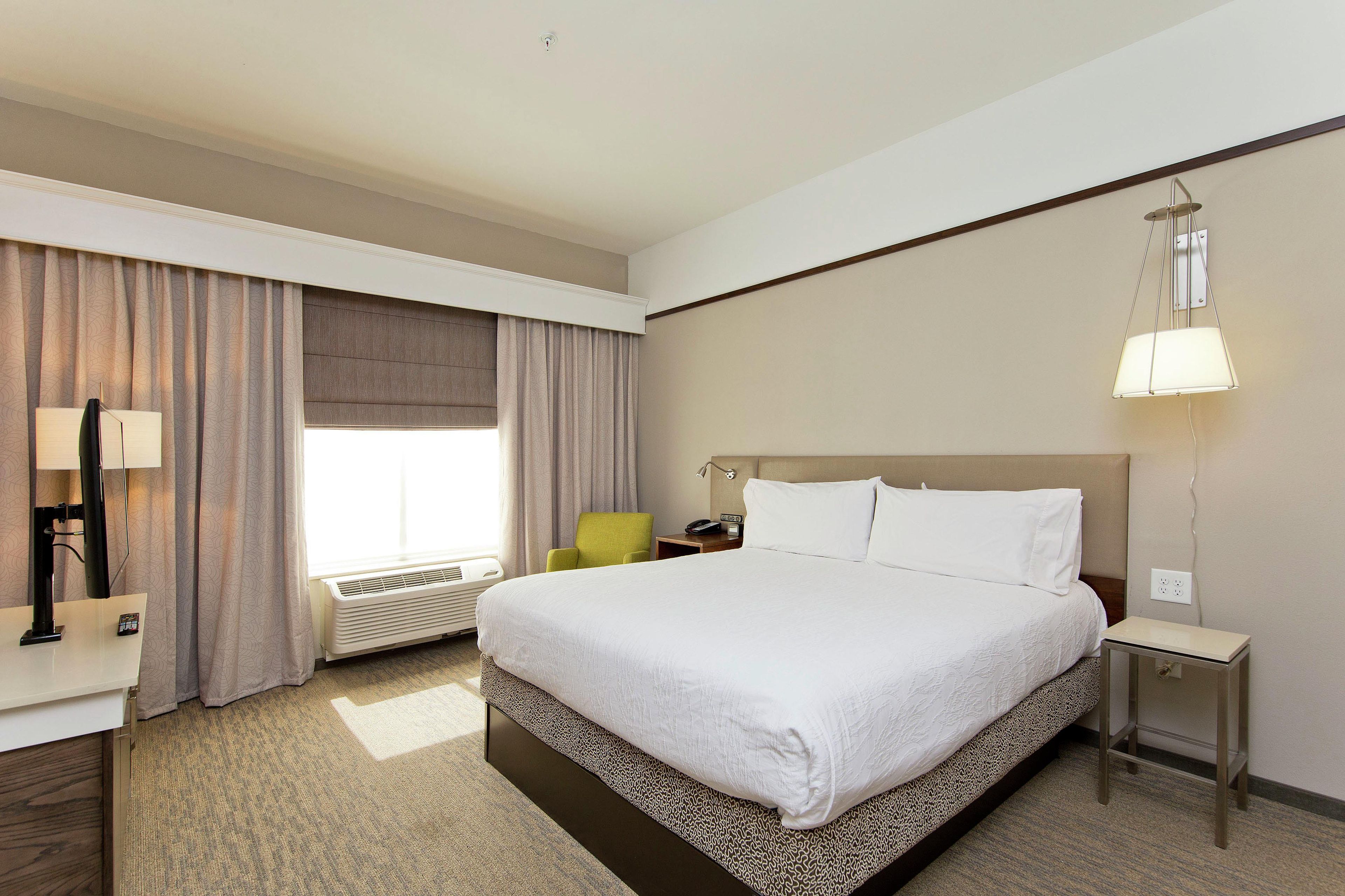 A hotel room with a large bed, a desk, a chair, a bedside table, a lamp, and a window with curtains.
