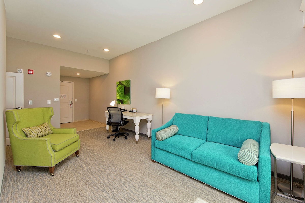 A room with a green armchair, a teal sofa, a desk with a chair, and a lamp.