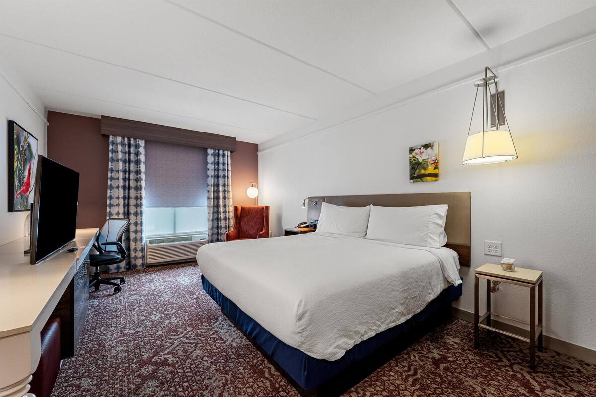 Ein Hotelzimmer mit einem großen Bett, einem Schreibtisch mit Stuhl, einem Fernseher, einer Lampe und einem kleinen Tisch mit einer Tasse.