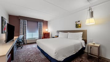 A hotel room with a large bed, a desk with a chair, a television, a lamp, and a small table with a cup.
