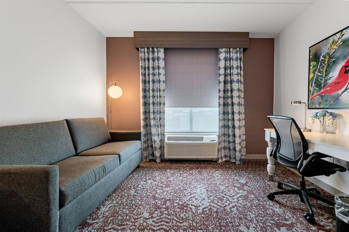 Ein Hotelzimmer mit einer Couch, einem Schreibtisch mit Stuhl, einem großen Fenster mit Vorhängen und einer Wandklimaanlage.
