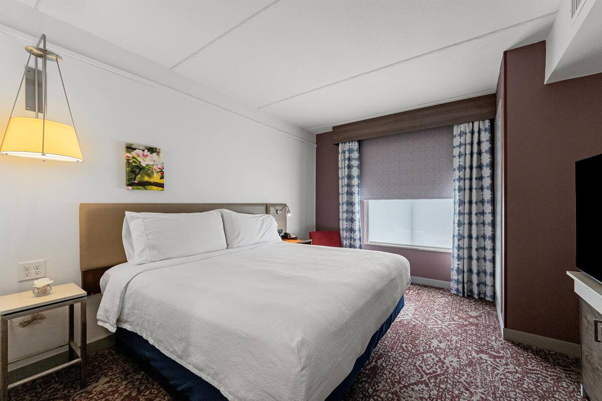 Ein Hotelzimmer mit Bett, Nachttisch, Lampe, Fernseher, einem Fenster mit Vorhängen und einem Bild an der Wand.
