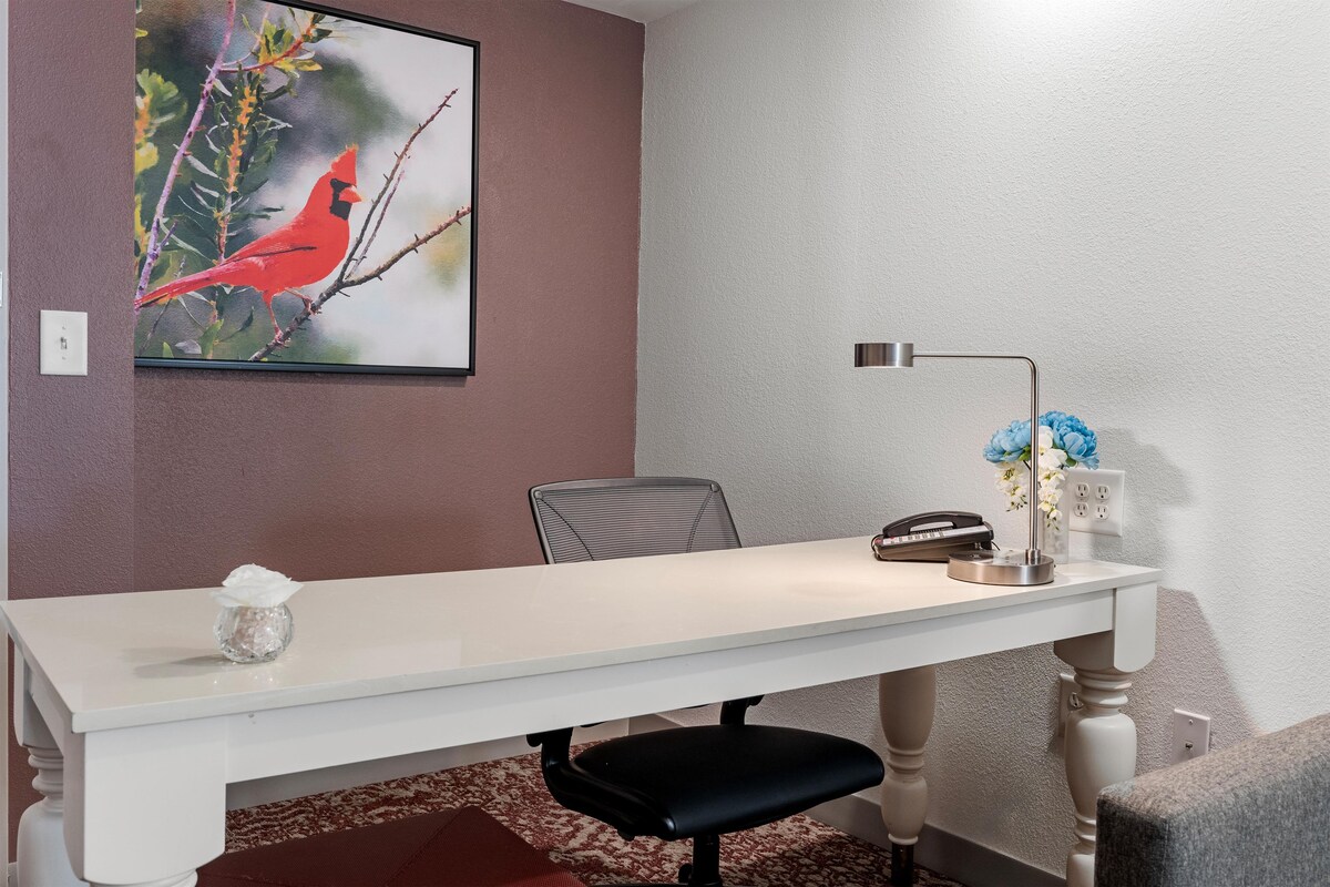 Una sala de conferencias con una mesa blanca, una sola silla de oficina negra, un cuadro enmarcado con una imagen de un ave, una lámpara de escritorio y un jarrón con flores.