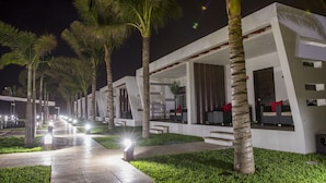 Un hotel moderno con un camino bordeado por palmeras, zonas de estar al aire libre e iluminación ambiental.
