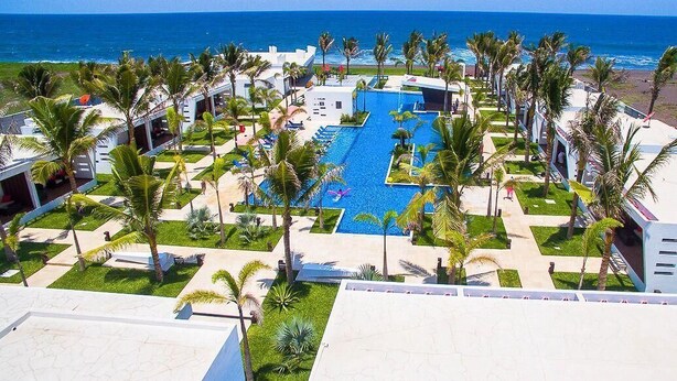 Un resort con piscina central rodeada de palmeras y vista al océano.