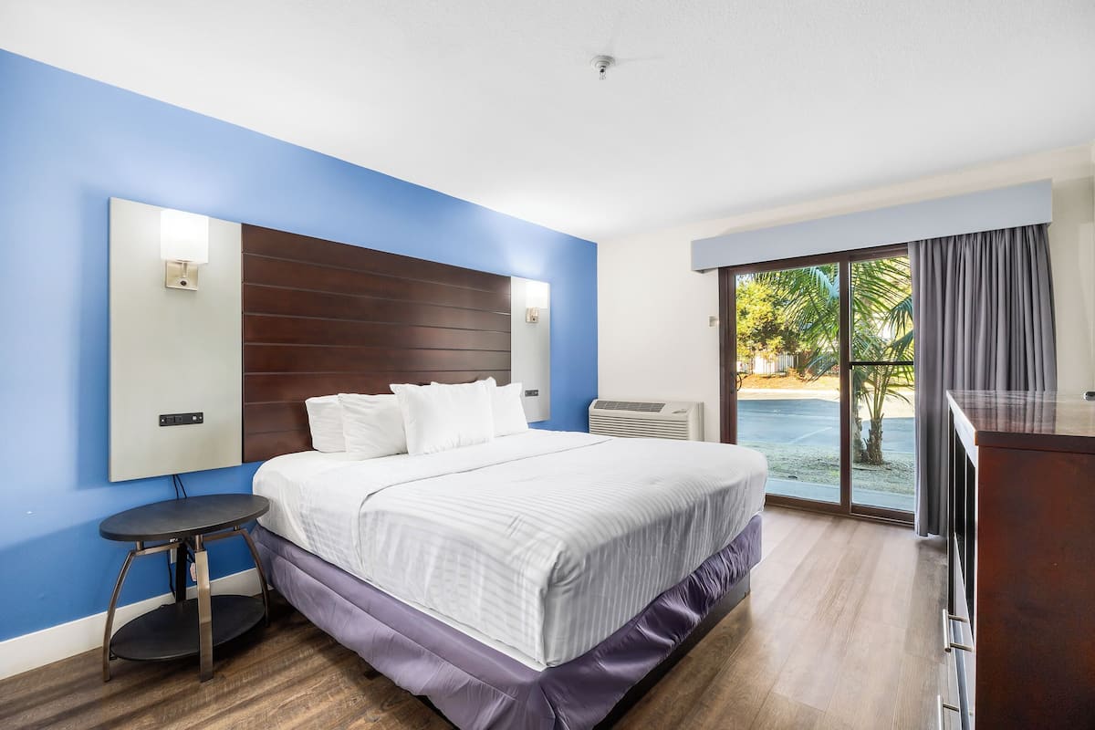 A hotel room with a bed, a small table, a mirror, and a view of the outdoors.