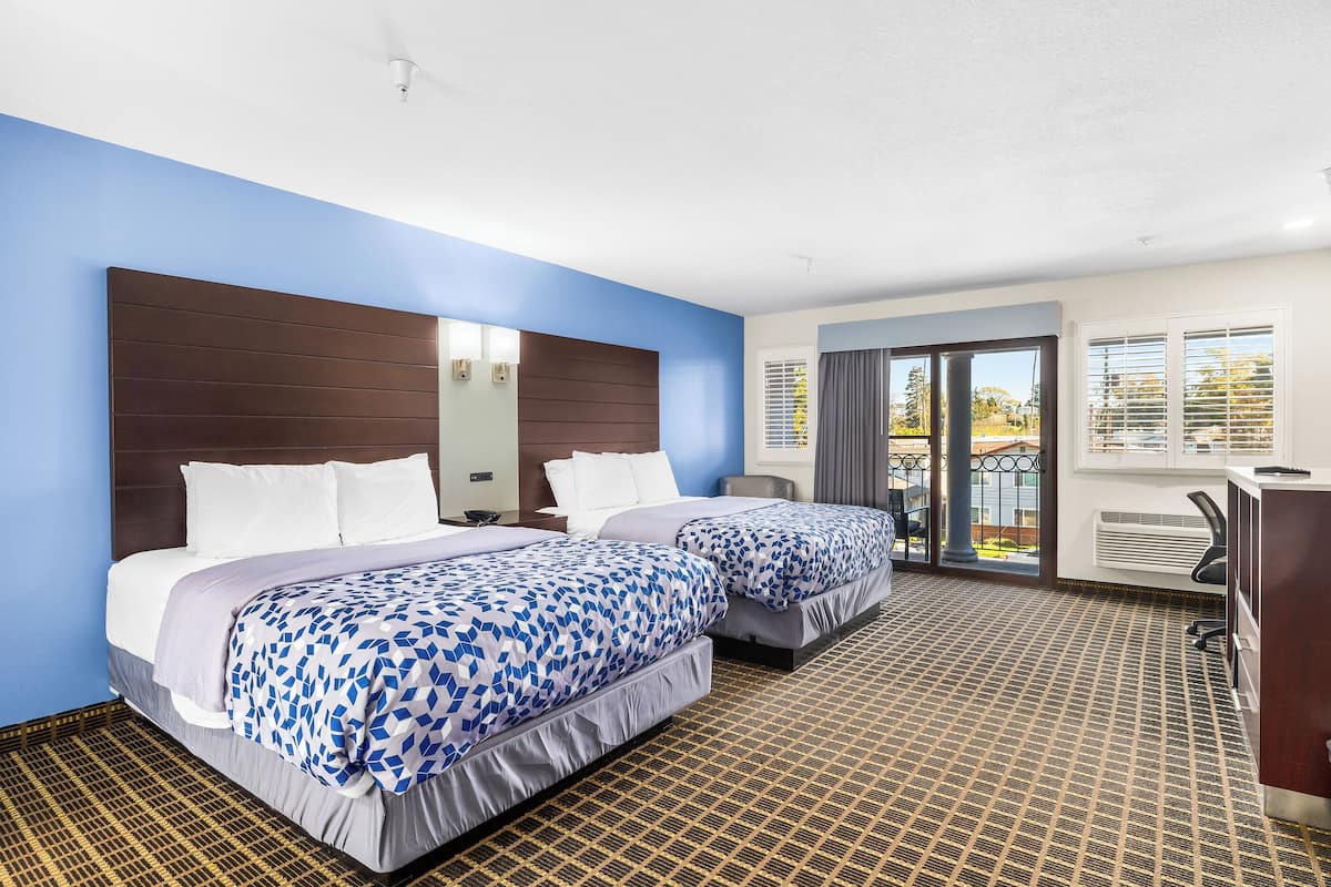 A hotel room with two beds, a large window with blinds, a desk, and a view of the outdoors.