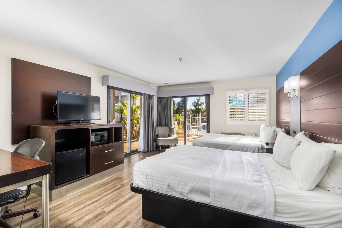 A hotel room with two beds, a desk with a TV, a mini-fridge, and a view of the outdoors.