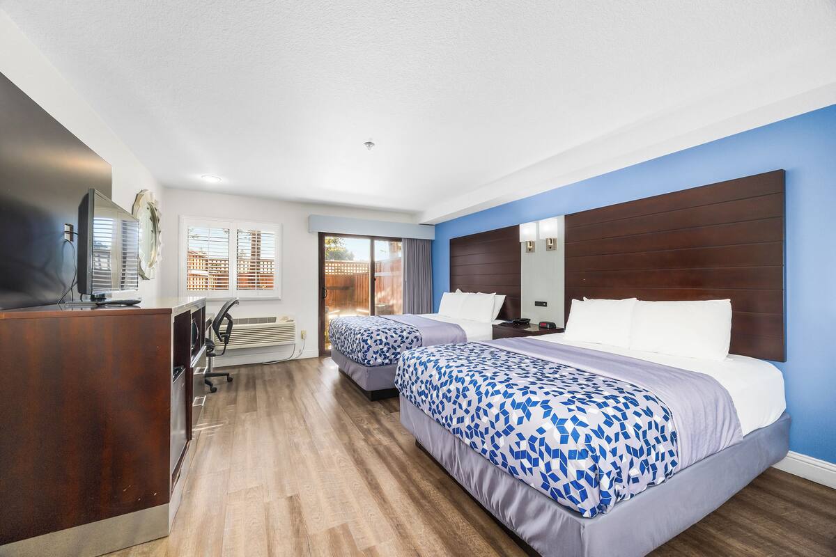 A hotel room with two beds, a desk, a TV, and a large window with blinds.