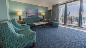 Une chambre d’hôtel avec un tapis bleu, un canapé, un fauteuil, une petite table, deux lampes, un tableau et un balcon avec vue.