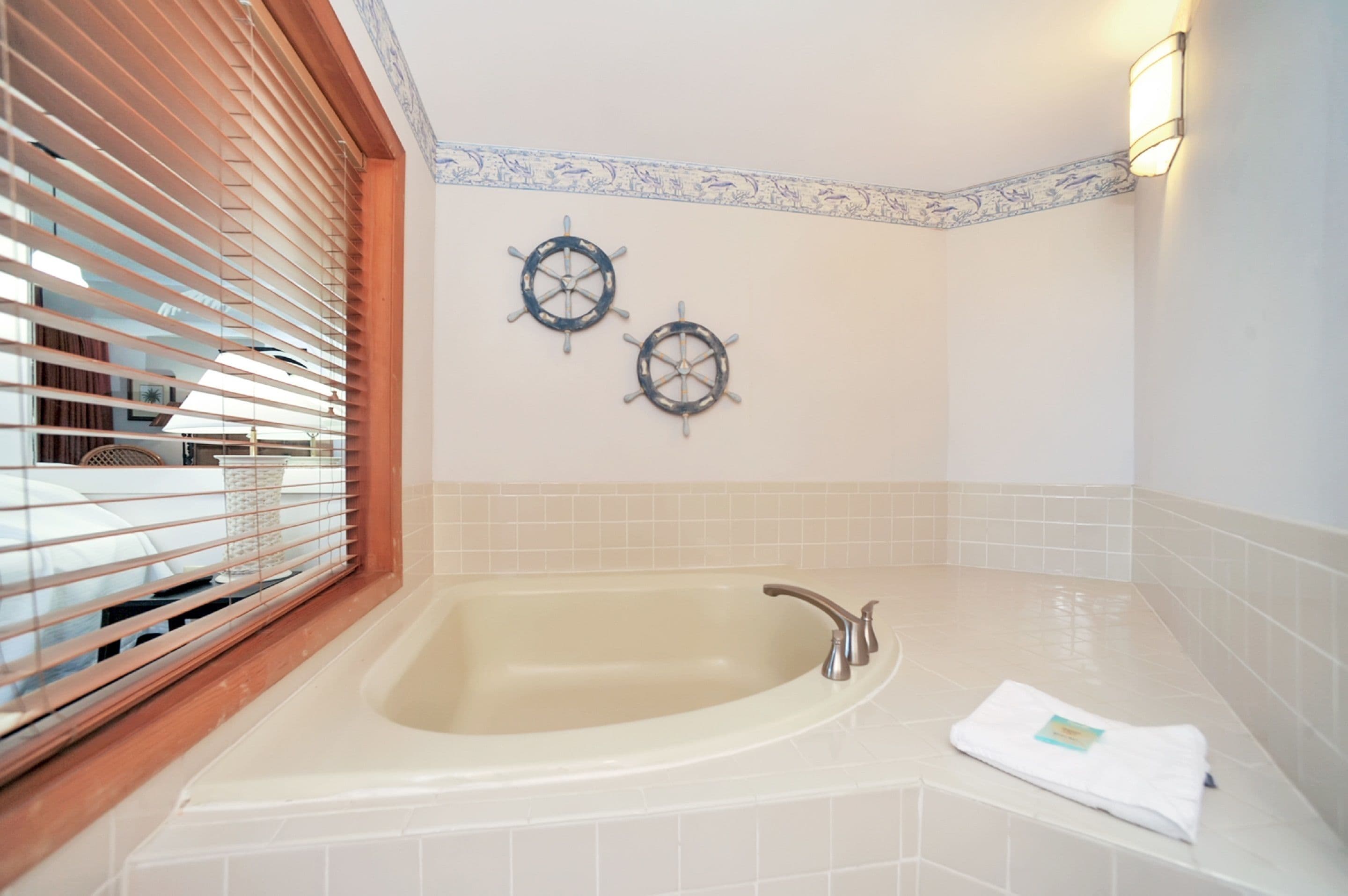 A bathroom with a bathtub, two ship wheel wall decorations, a towel on the edge, and a window with blinds.