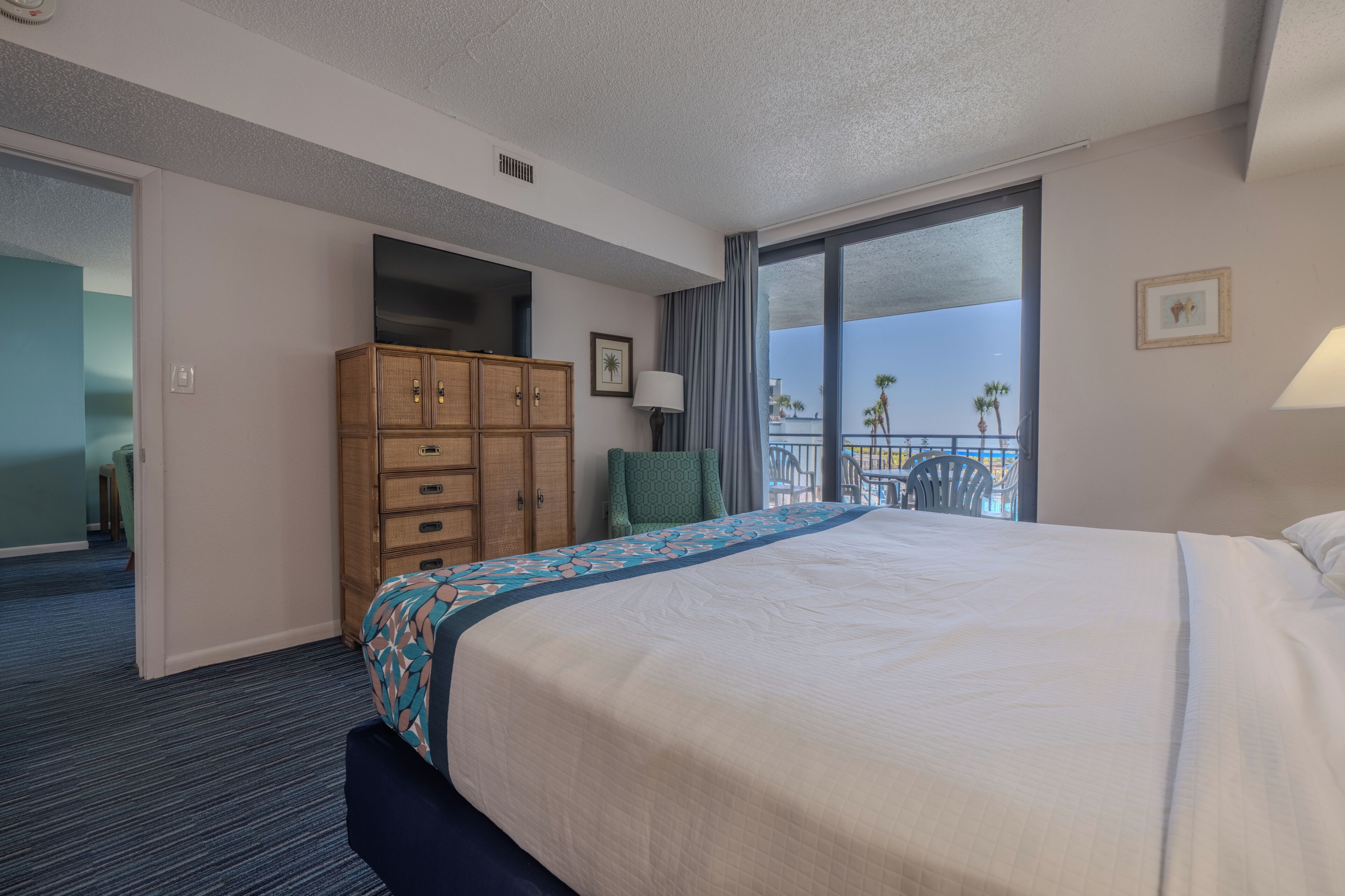 Une chambre d’hôtel avec un grand lit, une armoire en bois, une chaise, un balcon avec vue et une porte donnant sur une autre pièce.