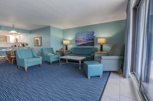 A living room with teal patterned furniture, a painting of a beach scene, and a view of the kitchen.