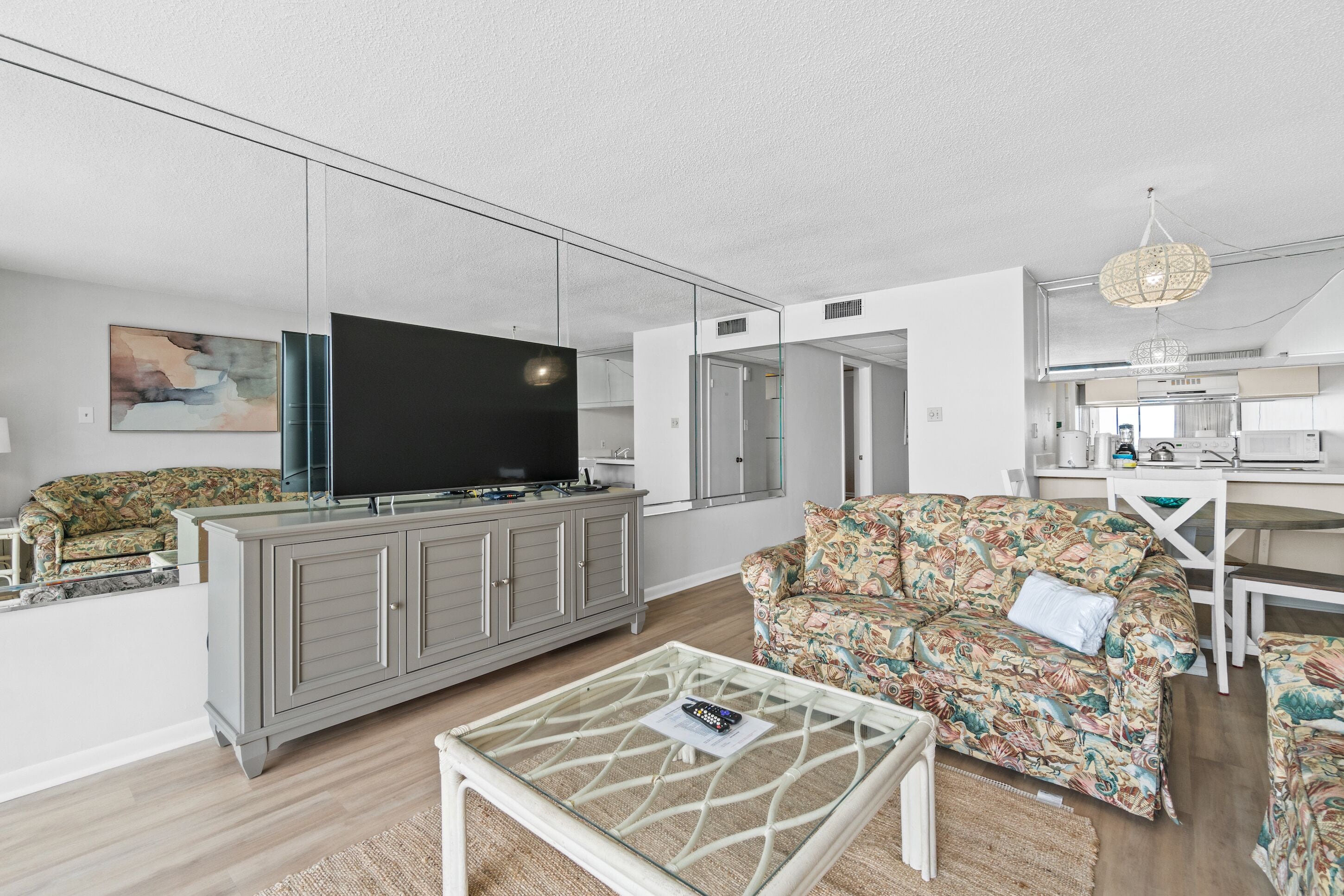 A modern living room with a flat-screen TV, a sofa, a coffee table, and a kitchen area visible through a glass partition.