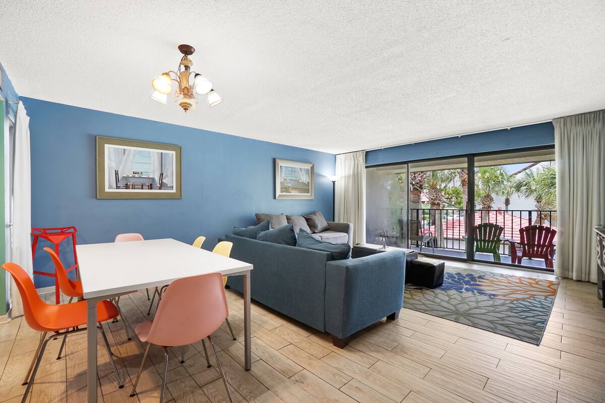 Un salon moderne avec un mur bleu, une table blanche, des chaises orange et roses, un canapé bleu et un balcon avec des chaises rouges.