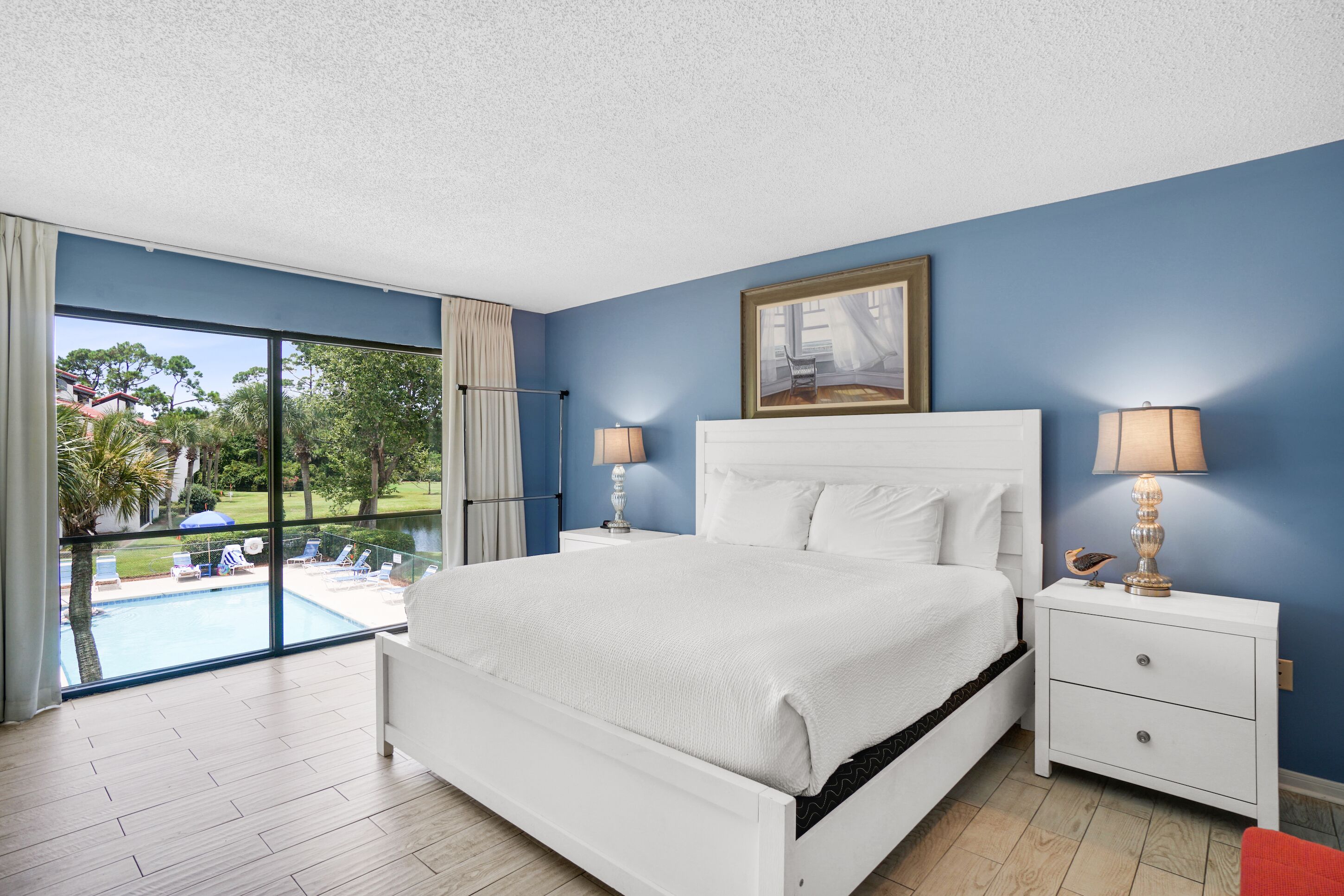 A bedroom with a large bed, two bedside tables with lamps, a painting above the bed, and a view of a swimming pool and greenery outside.