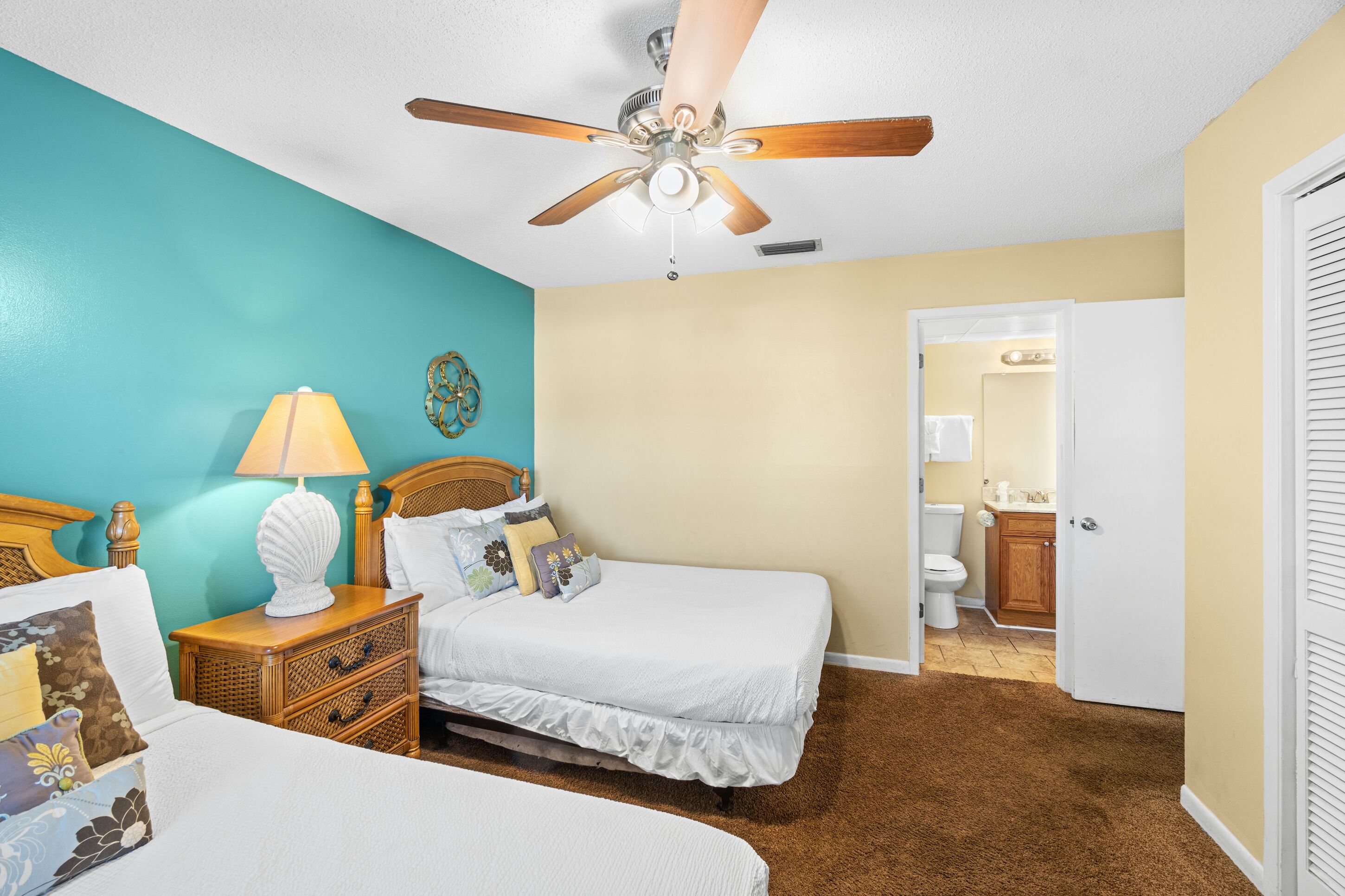 A bedroom with two beds, a ceiling fan, a nightstand with a lamp, and a bathroom visible through an open door.