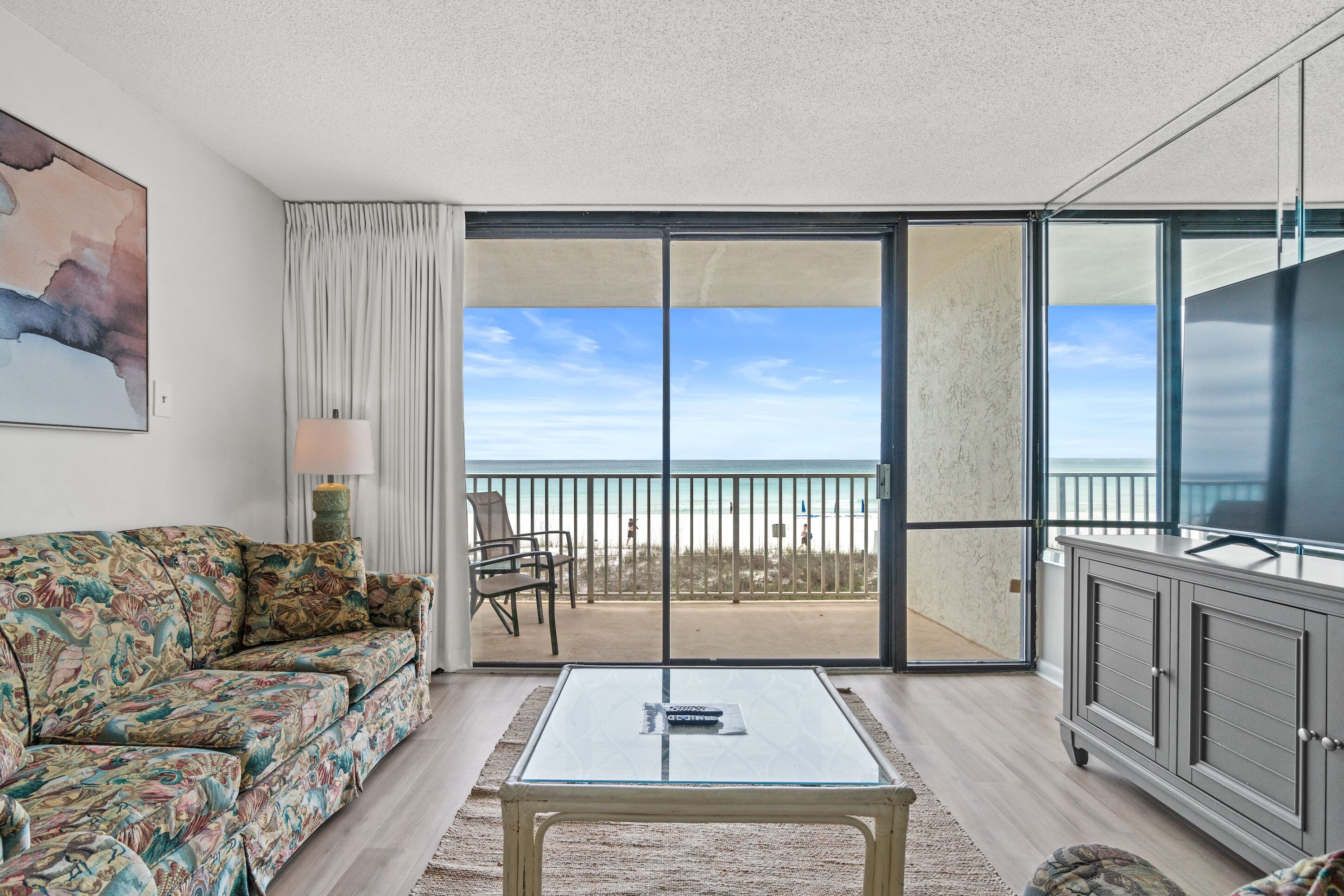 A living room with a sofa, a glass coffee table, a cabinet, and a view of the beach through a sliding glass door.