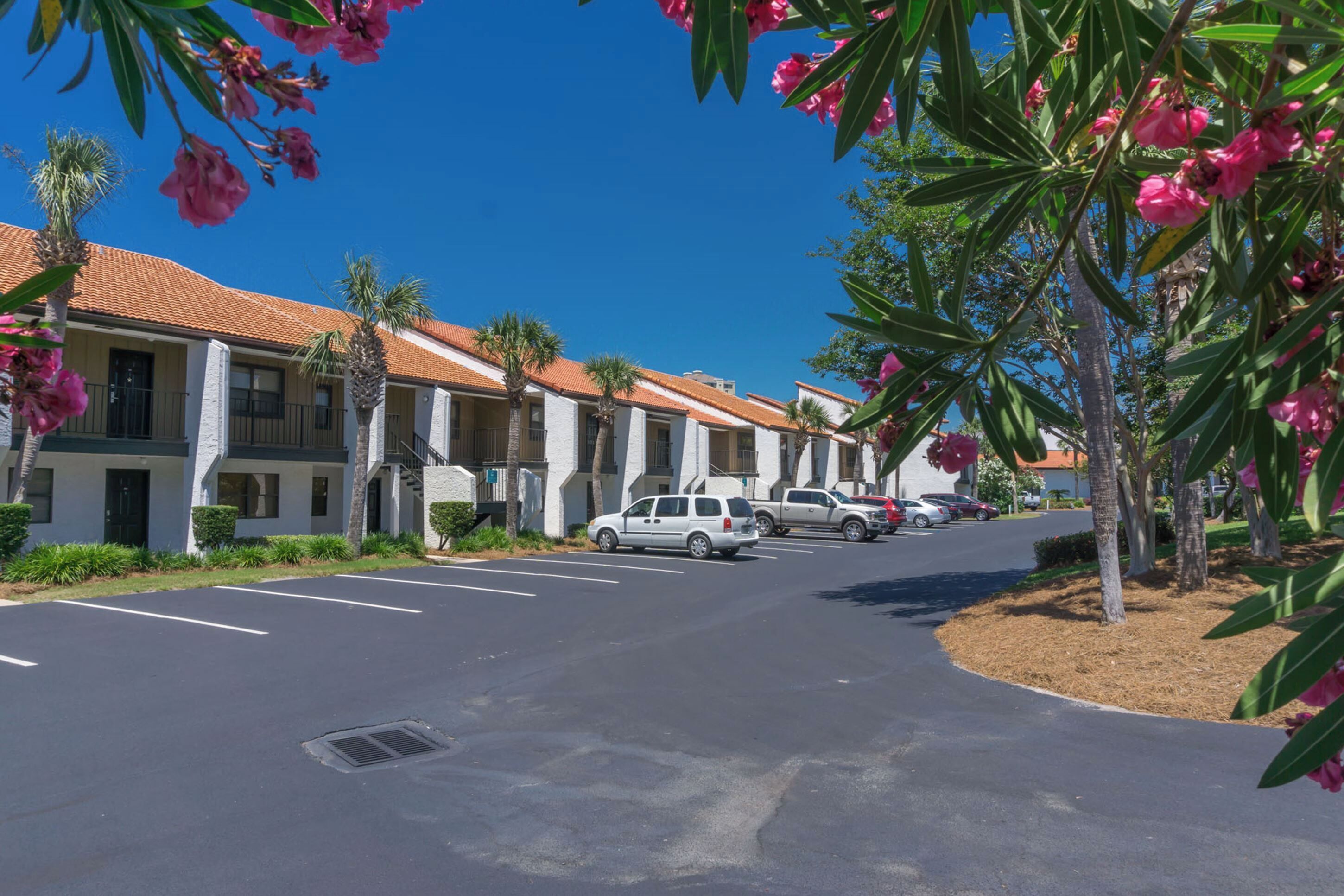 A row of white buildings with red-tiled roofs, parked cars, and palm trees.