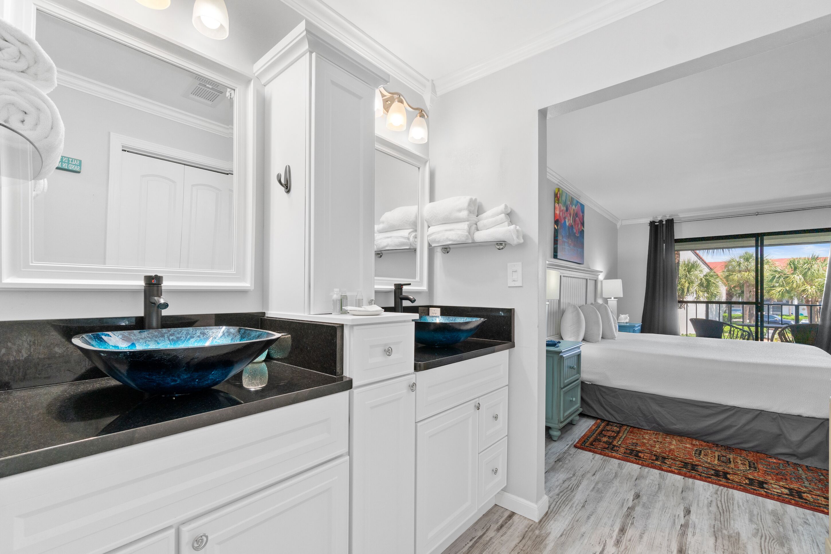 Une salle de bain moderne avec un grand miroir, des meubles blancs, un lavabo en verre bleu et un lit donnant sur des palmiers.