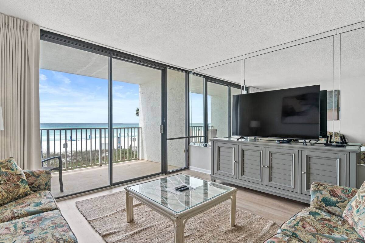 Un balcon spacieux avec une porte vitrée donnant sur la plage, un téléviseur à écran plat posé sur un meuble et une table basse en verre.