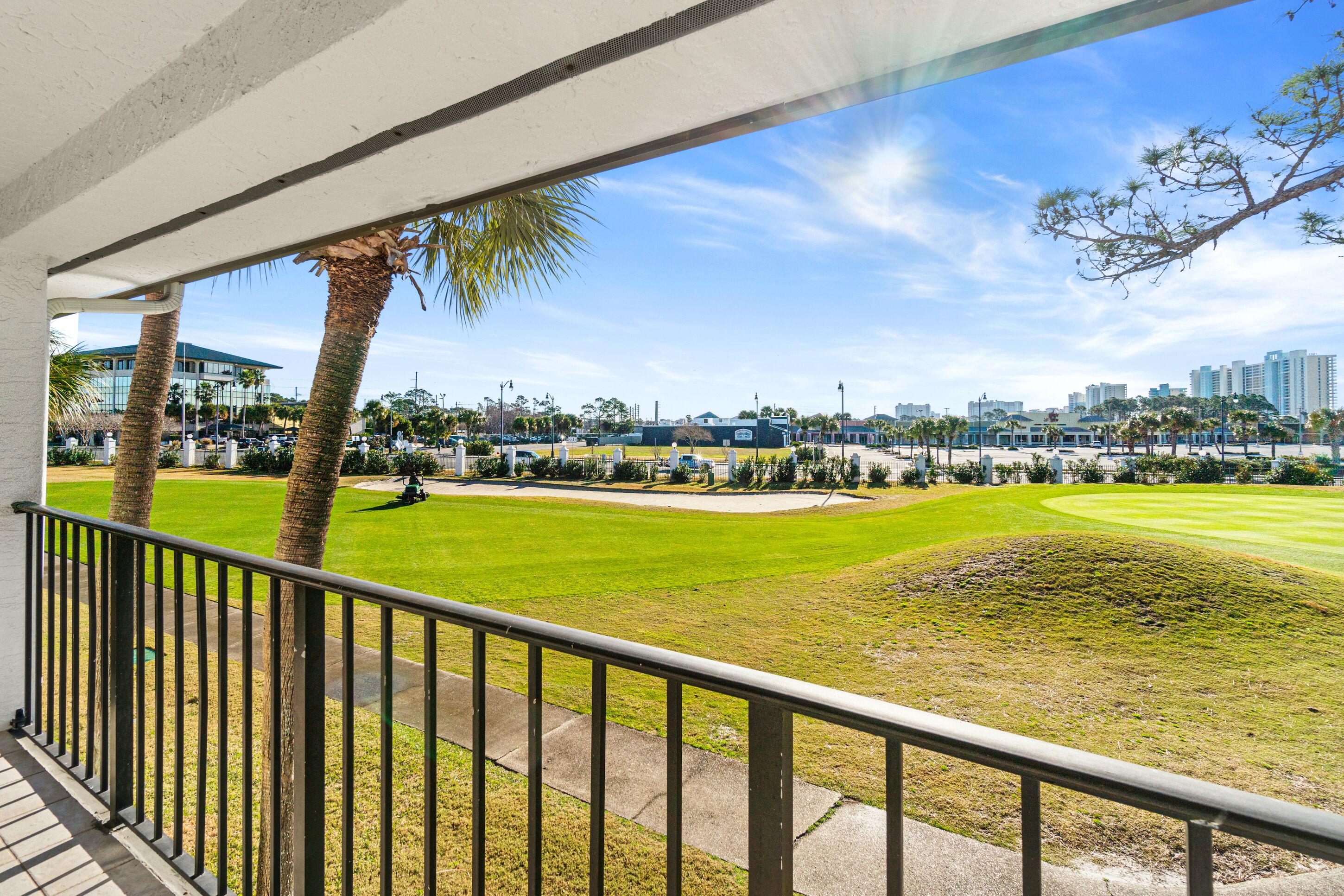 A view from a balcony overlooking a well-maintained lawn with a golf cart, palm trees, and a cityscape in the background.