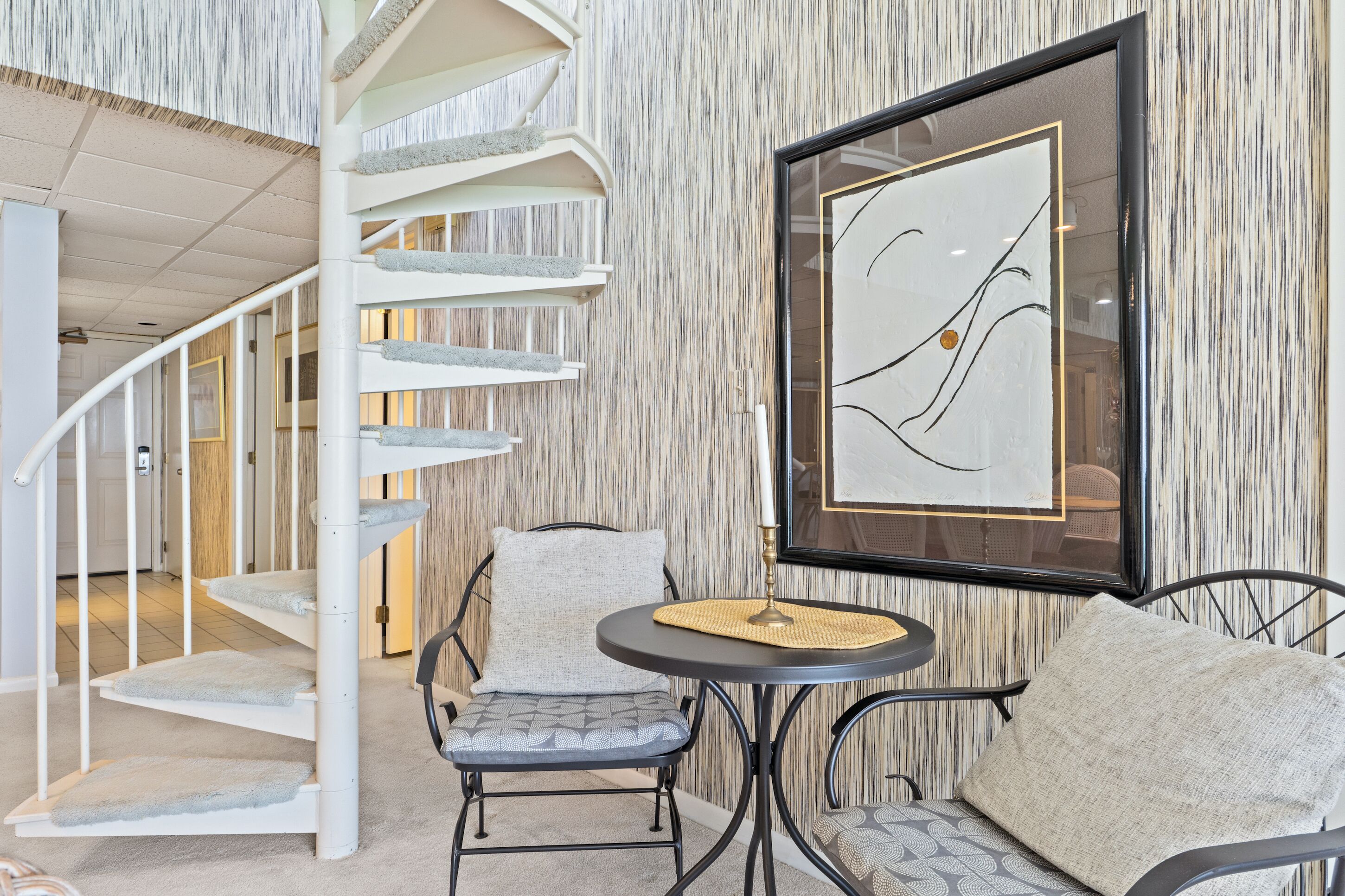 Un escalier en colimaçon au design moderne, une œuvre d’art abstraite encadrée et une table ronde avec deux chaises, le tout dans un espace intérieur chaleureux.