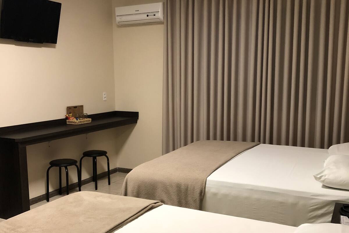 Een hotelkamer met twee bedden, een bureau met twee krukken, een flatscreen-televisie en een raam met gordijnen.