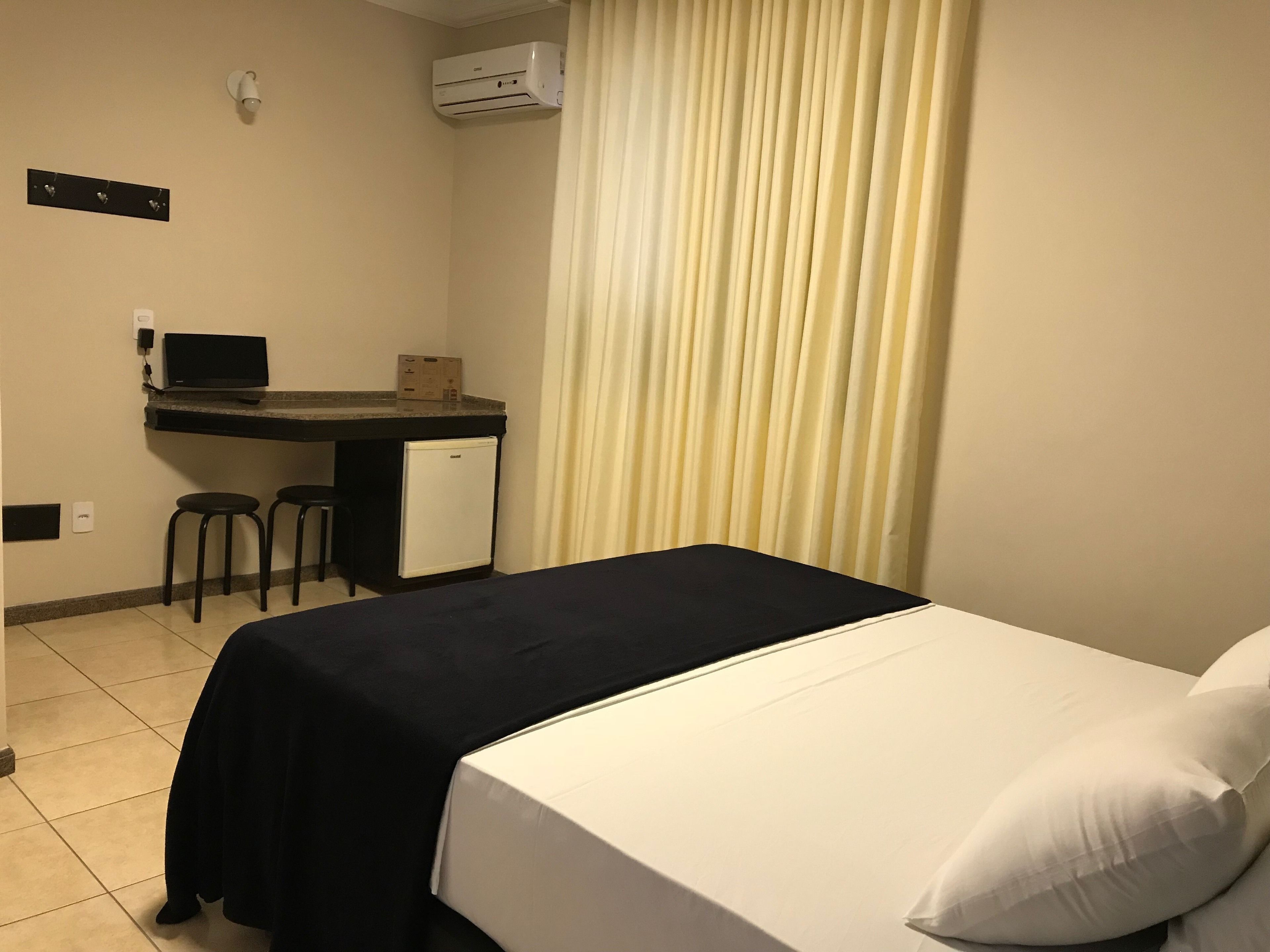 A hotel room with a bed, a desk with a computer, two chairs, and an air conditioning unit.
