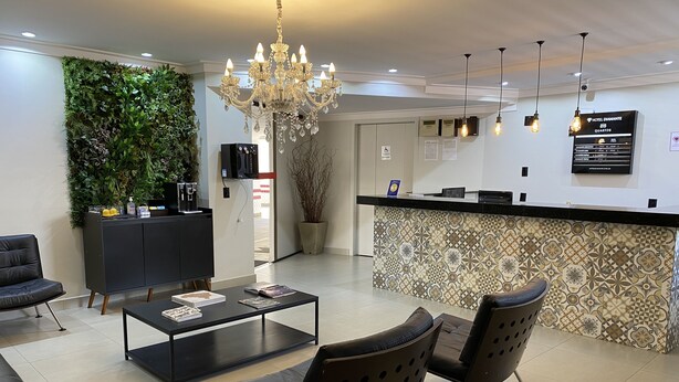 A hotel lobby with a reception desk, a chandelier, a coffee machine, a potted plant, and a seating area with two chairs. - Hotel Diamante (São Paulo)