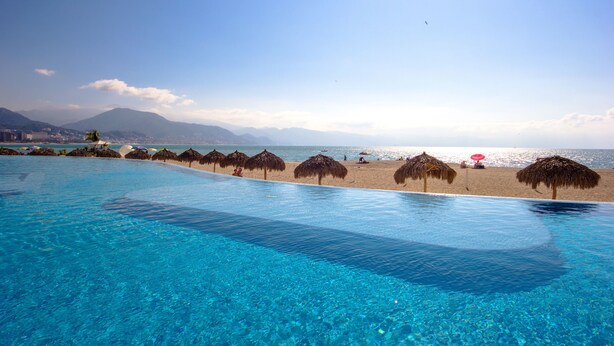 Ein klares, blaues Schwimmbecken mit Blick auf den Strand, Reetdachschirme und Berge im Hintergrund.