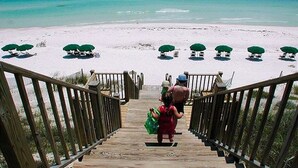 Un escalier en bois mène à une plage de sable blanc bordée d’une eau turquoise. On y trouve des parasols verts et des chaises longues.