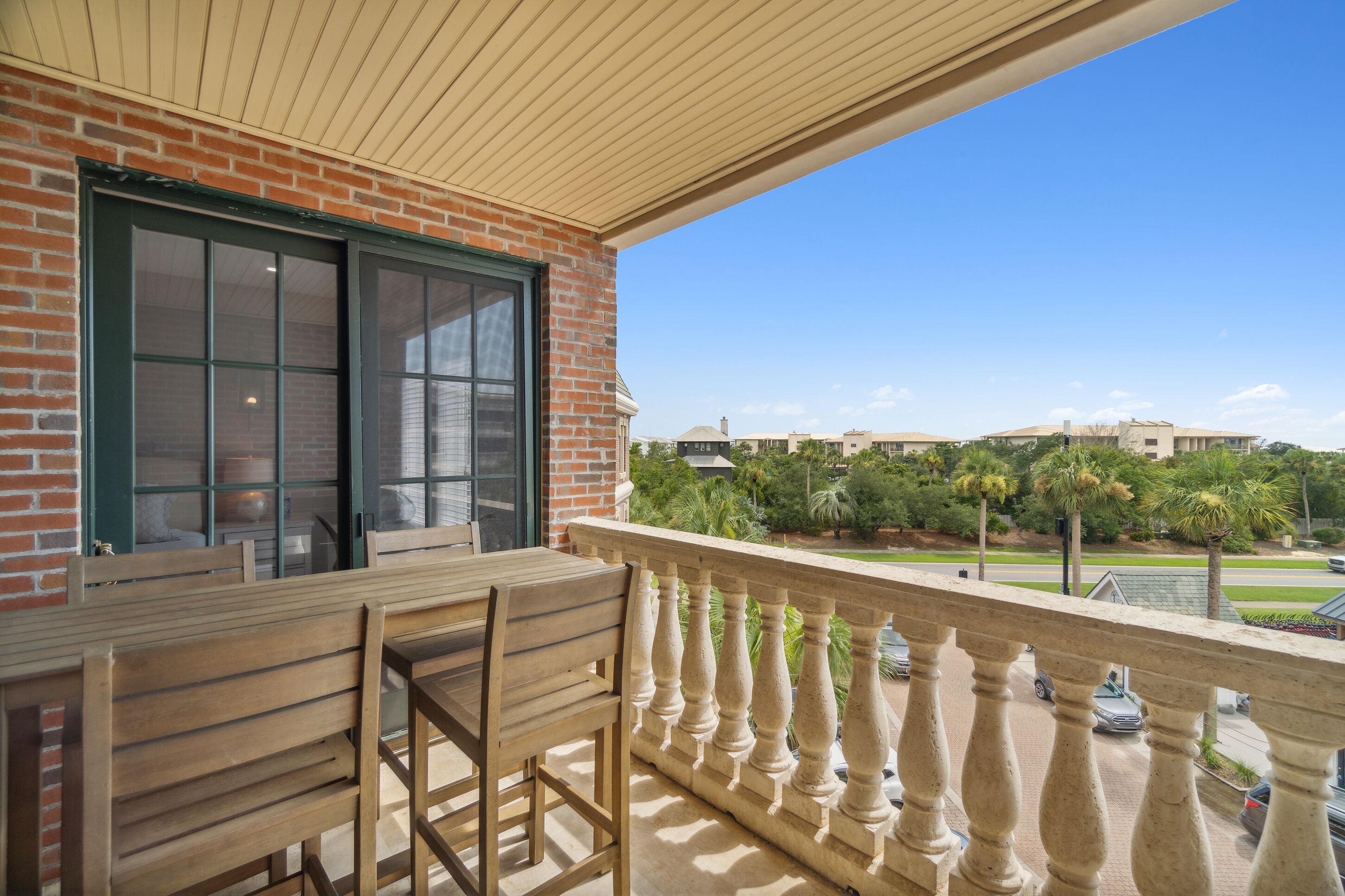 Un balcón con mesa y sillas de madera, un muro de ladrillo y vistas a una zona residencial con palmeras y casas.