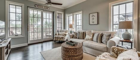 A spacious living room with a large window, a ceiling fan, and a sign reading "BEACH HOUSE".