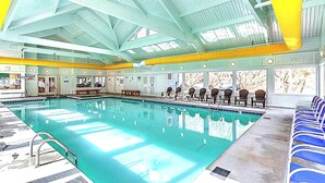 Una piscina interior con agua azul cristalina, rodeada de sillones de descanso y un techo alto con tragaluces.